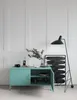 A teal metal cabinet with an open door, displaying items inside, stands against a white paneled wall. A black floor lamp and a framed abstract artwork are positioned to the right of the cabinet. Voir les images du produit