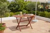 A wooden folding table set up on a patio with drinks and a bowl of fruit, surrounded by plants and a white umbrella. Produktbild-Vorschau