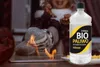 A person's feet in socks are near a fire, with a bottle of bioethanol in the foreground. Produktbild-Vorschau