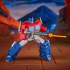 A red and blue robot toy, Optimus Prime, is posed in a dynamic stance, holding a rifle with an orange projectile. The background depicts a mountainous landscape with orange rock formations. Anteprima dell’immagine prodotto