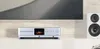 A silver audio system sits on a white surface in front of a light wood-paneled wall, with a large dark screen above and a white speaker to the right. Podgląd zdjęcia produktu