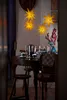 A table setting with a coffee pot, cups, candles, and a vase of flowers, illuminated by star-shaped lanterns. Produktbild-Vorschau