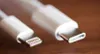 A close-up shot of two white charging cables, one with a Lightning connector and the other with a USB-C connector, lying on a wooden surface. Produktbild-Vorschau