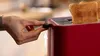 A hand is shown adjusting the lever of a red toaster with toast inside. Voir les images du produit