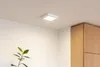 A square white LED ceiling light is mounted on a white ceiling in a room with white walls and wooden cabinets. Náhľad obrázku produktu