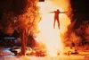 A man is engulfed in a large explosion, arms outstretched, appearing to fly upwards. Several cars are visible in the foreground, and houses and trees are in the background. Podgląd zdjęcia produktu