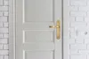 A close-up of a white door with a gold handle, next to a white brick wall with an electrical outlet. Anteprima dell’immagine prodotto