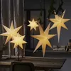Several illuminated paper star lanterns are displayed on a wooden table and hanging in a room with a curtained window. Podgląd zdjęcia produktu