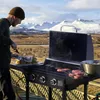 A man grilling burgers outdoors with a snowy mountain range in the background. Produktbild-Vorschau