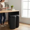 A woman is standing in a kitchen next to a counter, with two black trash bins underneath. Voir les images du produit