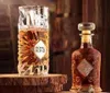 A bottle and a glass of amber liquid, likely a beverage or perfume, are displayed on a wooden surface with a blurred background. Podgląd zdjęcia produktu