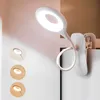 A white clip-on lamp with a ring-shaped light is attached to a white surface. Produktbild-Vorschau