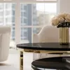 A close-up of a round coffee table with a black top and gold trim, featuring a vase of cream-colored roses. The background shows a bright room with large windows and upholstered chairs. Náhled obrázku produktu