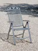 A metal folding chair sits on a gravel surface, with dappled sunlight and shadows from trees overhead. Produktbild-Vorschau