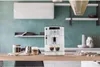 A white espresso machine is on a wooden table in a modern kitchen setting. Produktbild-Vorschau