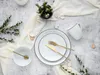 A flat lay of a white dinnerware set with black trim, gold cutlery, and a small vase of greenery on a textured gray background. Náhľad obrázku produktu