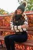 A woman wearing a green hat and scarf sits on a wooden bench in front of red foliage. Voir les images du produit