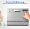 A close-up of a silver countertop dishwasher with a hand pressing a button on its control panel. Kitchen utensils and a teacup are visible on the left side. Produktbild-Vorschau