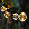 A string of decorative lights with clear, round bulbs, illuminated and hanging outdoors amongst foliage. Náhľad obrázku produktu