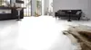 A modern living room and kitchen area with white flooring, a dark sofa, a kitchen island with a stool, and a rug. Produktbild-Vorschau
