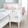 A white crib is attached to a larger bed, with a pink bunny toy and blankets on the bed. Produktbild-Vorschau