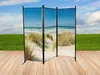 A three-panel room divider with a beach scene printed on it, set up on a wooden deck with a grassy field and sky in the background. Produktbild-Vorschau
