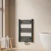 A dark grey towel radiator mounted on a light beige wall, next to a window and a wooden stool with a folded towel. Produktbild-Vorschau