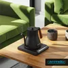 A black electric kettle with a wooden handle sits on a wooden table next to two cups, with green sofas in the background. Náhled obrázku produktu