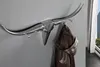 A silver coat rack shaped like a bull's head is mounted on a gray wall, with a brown jacket hanging on one of the hooks. A window with buildings visible can be seen in the background. Produktbild-Vorschau