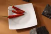 A white square plate with three red chili peppers on it, placed on a brown surface with dark rocks scattered around. Náhled obrázku produktu