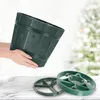 A person holds a green Christmas tree stand, with two green tree stand bases in the foreground, and a blurred Christmas tree in the background. Voir les images du produit