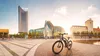 A bicycle is parked in a modern city square with contemporary buildings and a fountain in the background during sunset. Produktbild-Vorschau