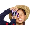 A young girl wearing a straw hat and a blue sweater with red heart details is holding a purple toy camera to her eye and smiling. Voir les images du produit