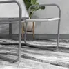 A close-up view of a modern grey metal chair with white mesh seating, placed on a patterned rug in front of a concrete wall and a potted plant. Náhled obrázku produktu