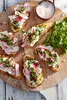 A wooden board with several bruschetta topped with cream cheese, radishes, and herbs, a small bowl of salt, and a pile of greens. Podgląd zdjęcia produktu