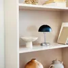 A black and chrome table lamp sits on a shelf next to a decorative bowl. Produktbild-Vorschau