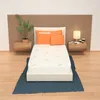 A mattress is displayed on a bed frame, with pillows and a bedside table with a lamp and cups. Produktbild-Vorschau