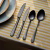 A set of four dark gray cutlery pieces (two knives and two spoons) are laid out on a striped yellow and white tablecloth, with a portion of a blue and tan plate visible on the left. Podgląd zdjęcia produktu
