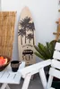 A surfboard with a California surf trip design leans against a white wall, next to a bamboo screen and a white chair with a bowl of fruit and a mug on a small table. Náhled obrázku produktu