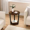A round side table with two shelves is placed between two armchairs in a living room setting. A vase with flowers and a cup of coffee are on the top shelf, and a book is on the bottom shelf. Produktbild-Vorschau