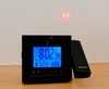 A black digital alarm clock with a blue display is shown on a wooden surface. The clock projects the time onto the wall behind it. Produktbild-Vorschau