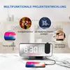 A digital alarm clock with a projection feature, shown on a marble surface with a smartphone charging next to it. The image highlights various features of the clock. Produktbild-Vorschau