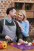 A couple is in a kitchen preparing food. The man is wearing an apron and chopping vegetables on a cutting board, while the woman stands behind him, resting her hand on his shoulder and smiling. Náhled obrázku produktu