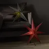 Two star-shaped paper lanterns are displayed in a dimly lit room, one dark green on a sofa and one red on the floor. Podgląd zdjęcia produktu