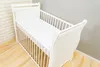 A white baby crib with a patterned sheet is shown in a room with a brick wall and a gray wall. Produktbild-Vorschau