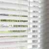 Close-up view of white horizontal blinds partially covering a window with greenery visible outside. Produktbild-Vorschau