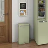 A green retro-style pedal bin is placed next to a green retro-style refrigerator, with a wooden door and a wall-mounted notice board in the background. Podgląd zdjęcia produktu