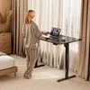 A woman stands at a standing desk in a bedroom, working on a laptop. The desk has an electric height adjustment and a control panel. Anteprima dell’immagine prodotto