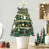 A decorated mini Christmas tree in a white pot, placed on a wooden surface with pinecones and a red ball nearby. In the background, there are blurred lights and other decorative Christmas trees. Náhľad obrázku produktu