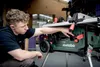 A young man is adjusting a green Metabo table saw in a workshop setting. Voir les images du produit